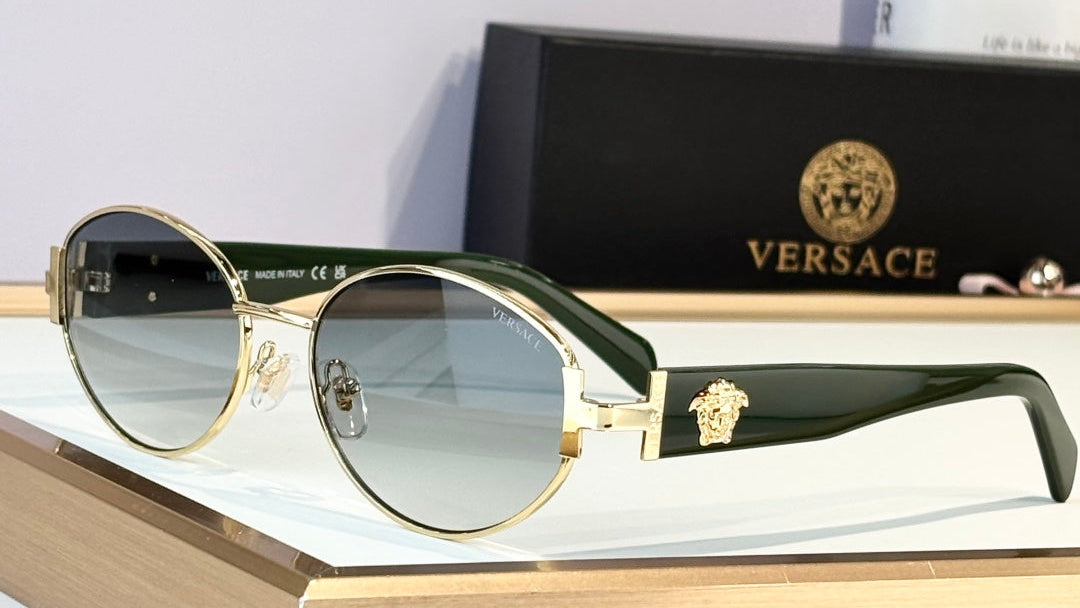 Versace oval sunglasses with gold metal frames, dark green temples, and a signature Medusa logo, displayed on a wooden block with the black branded box in the background.